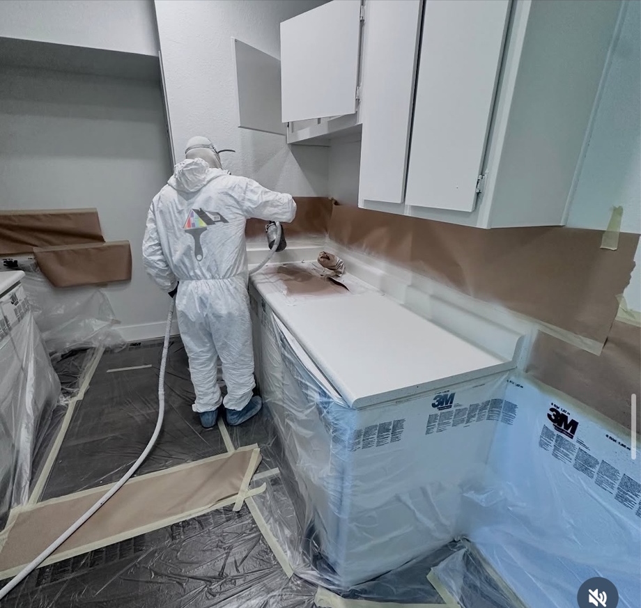 All In One Painting and Resurfacing crew member in full protective gear spray-refinishing a kitchen counter and cabinets